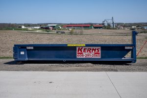 A large blue industrial dumpster with a sign that reads "Kerns" and a phone number is situated on a gravel area in front of a vast field near Madison, WI. Industrial buildings and structures are visible in the background under a clear blue sky.